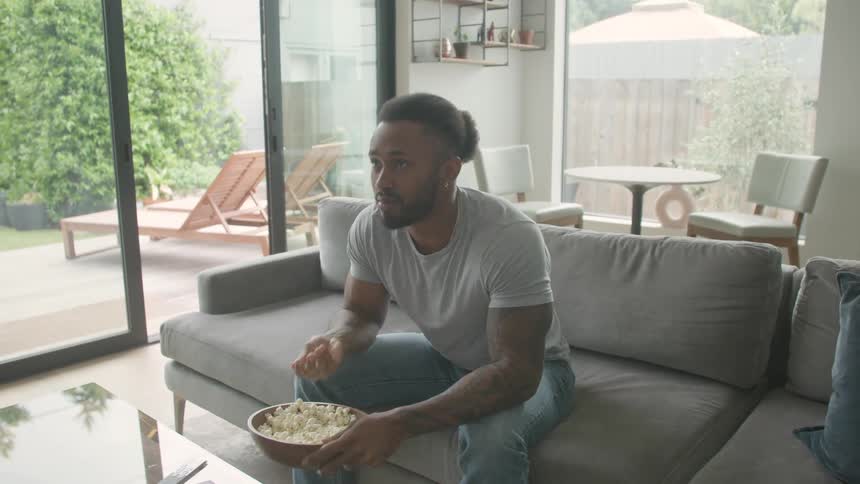 A young black man eating popcorn while watching tv in a living room during daytime