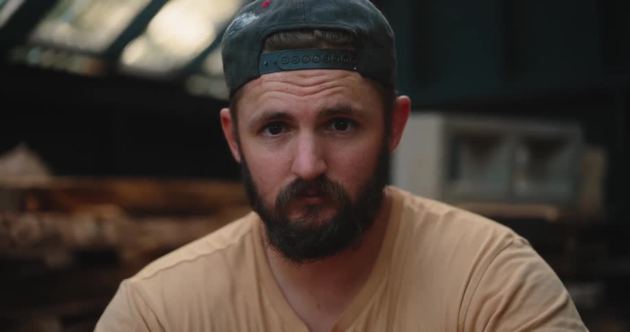 The video is a closeup of a bearded man wearing a tan tshirt and a dark baseball cap with red accents he is looking down intently and appears to be concentrating on a task out of frame possibly woodworking or sanding