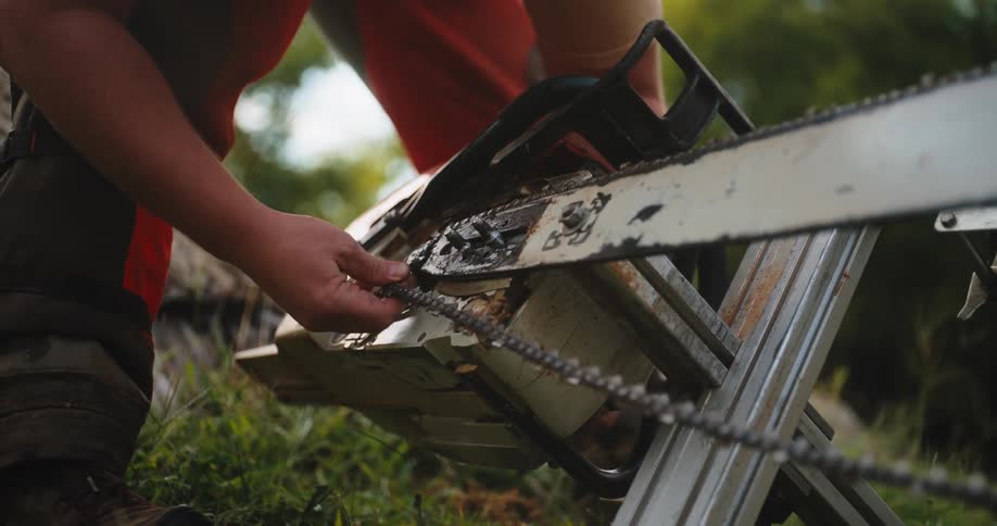 A close view of a worker in orange chaps adjusts the cutting chain on the long bar of a large chainsaw which is mounted to a metal frame outdoors on green grass