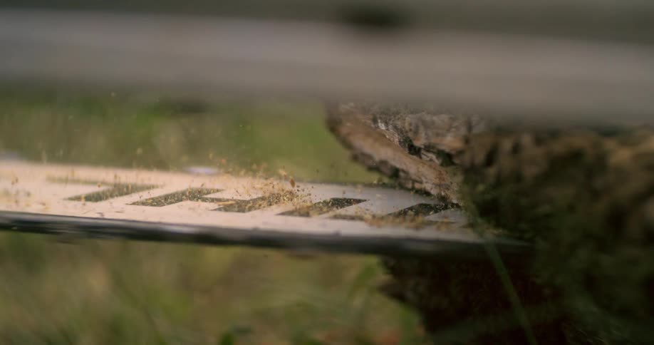 A closeup shot focuses on the metal guide bar of a running chainsaw as it cuts into a brown log lying on the ground causing small wood particles to fly upward and around the bar