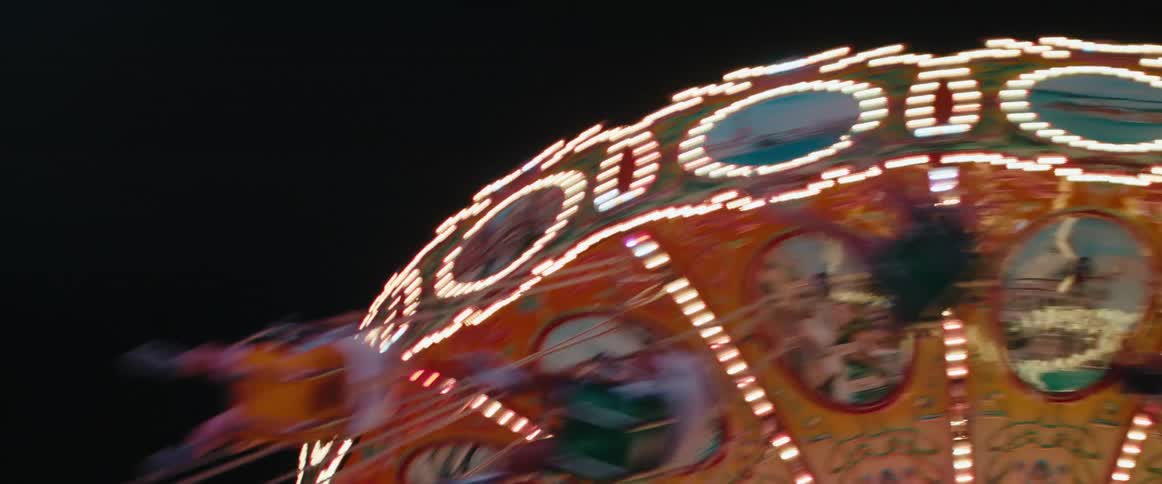 A brightly lit carnival swing ride spins at night its ornate panels and streaking lights blurring against the dark sky
