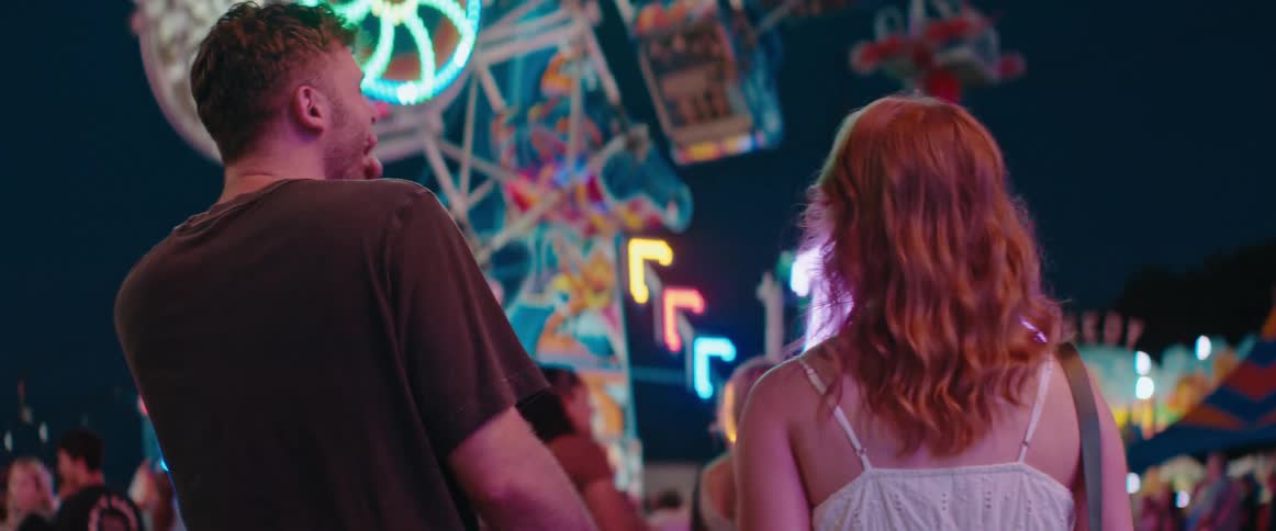 A man and a woman stand close together with their backs mostly to the camera then turn their heads to face each other with the man laughing widely against a dark background illuminated by many brightly colored neon lights and amusement park rides