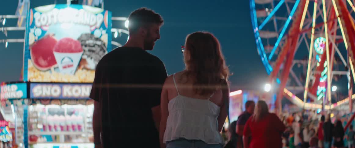 A man and a woman walk away from the camera holding hands and smiling at each other as they pass brightly lit carnival game booths and a ferris wheel at night