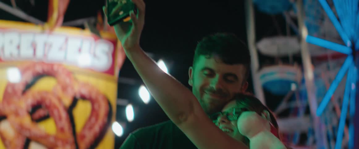 A happy woman wearing glasses holds up a smartphone to capture a selfie video while embracing a man who smiles and holds a prize stuffed animal at a colorful bright carnival at night