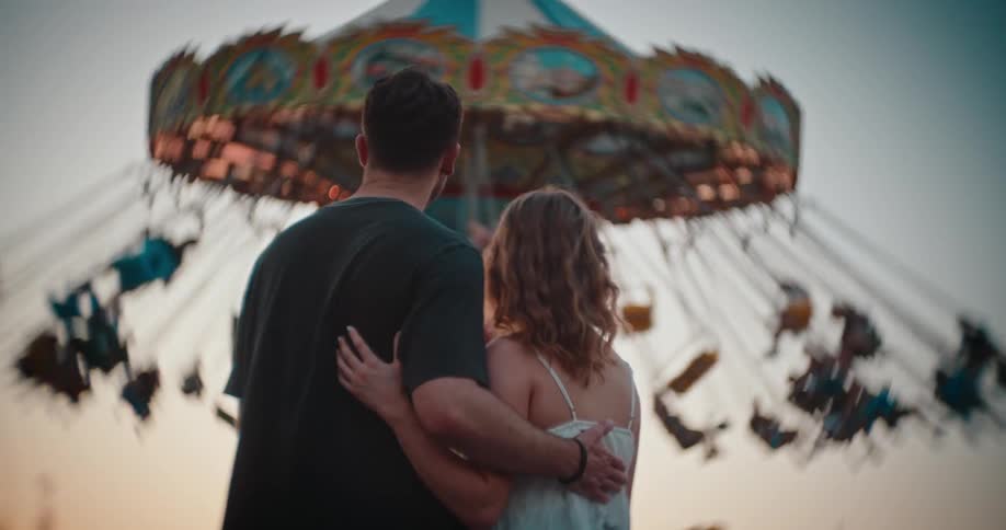 A man and a woman stand arm in arm watching a colorful swing ride spin at dusk