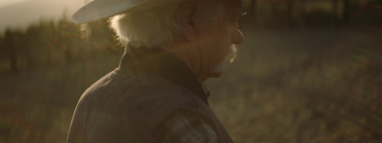 An elderly man in a cowboy hat walks outside in a rural area