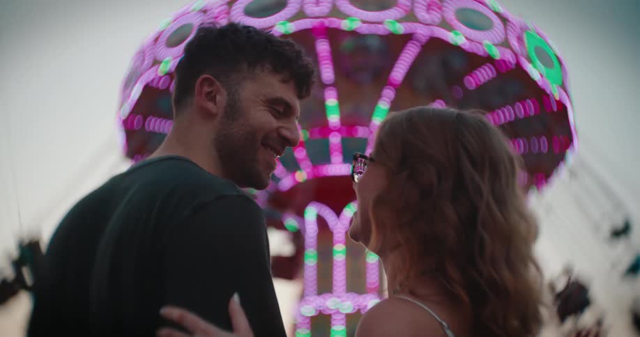 Couple leans in for a kiss in front of a brightly lit ride at the fair