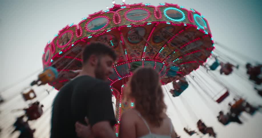 A man and a woman stand close to each other with their backs mostly to the camera looking up at a brightly lit rotating swing ride with red and blue lights while blurred riders swinging against a pale sky
