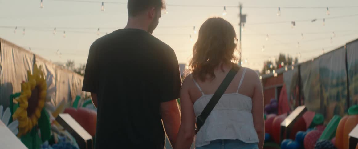 A rear view of a man and a woman holding hands and walking away from the camera into the intense lens flare of the setting sun surrounded by string lights and outdoor fair booths