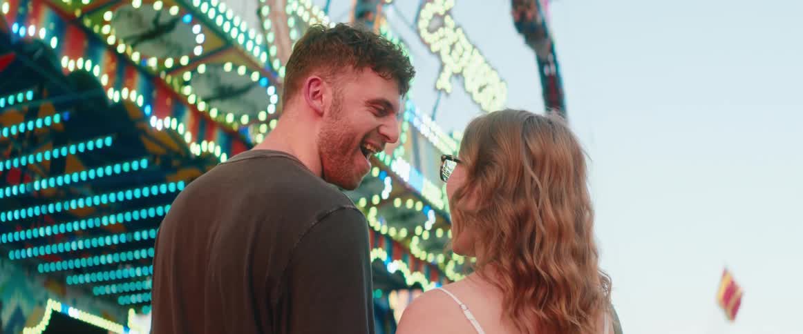 A man and a woman stand close to each other initially holding hands and then turn their heads to speak and laugh in an outdoor setting with a large brightly lit amusement park ride spinning in the background