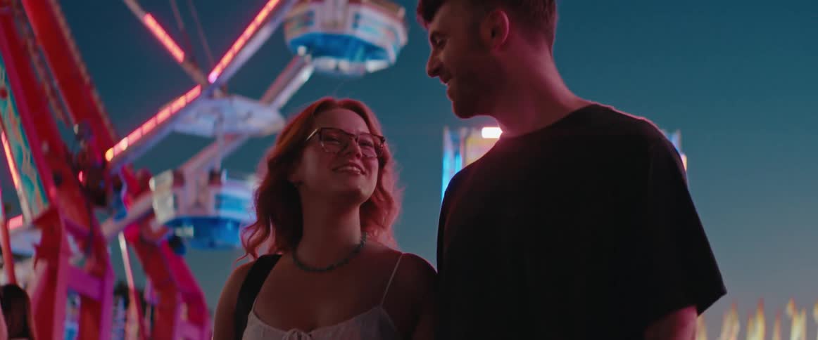 A happy young couple laughs and talks together at a carnival at night with a brightly lit ferris wheel in the background