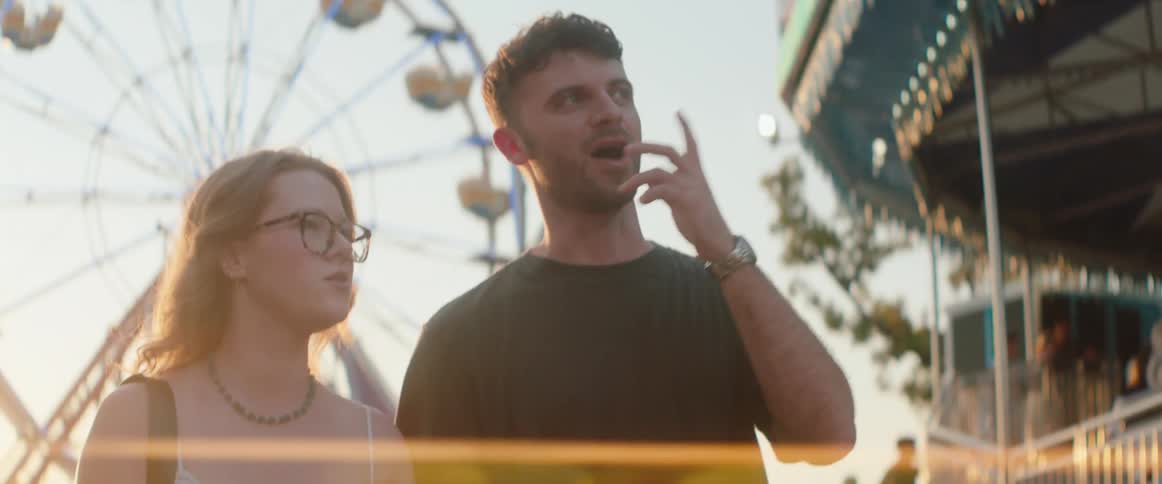 A man and a woman with eyeglasses stand outdoors at an amusement park featuring a large rotating wheel ride in the background