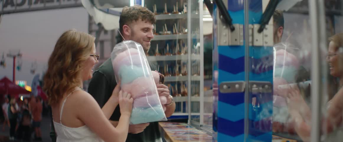 A smiling man and a woman stand at a carnival game booth with the woman holding a bag of pastel cotton candy and a pink unicorn prize as the man interacts with the booths attendant