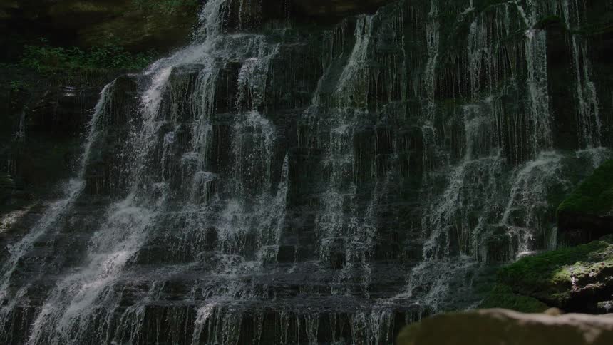 Sunlight filters through the trees illuminating a wide cascading waterfall as it flows down a dark layered rock face