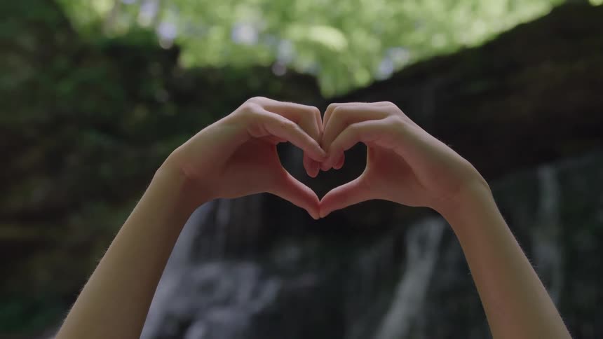 A persons hands form a heart shape framing a blurry waterfall and lush green foliage in the background