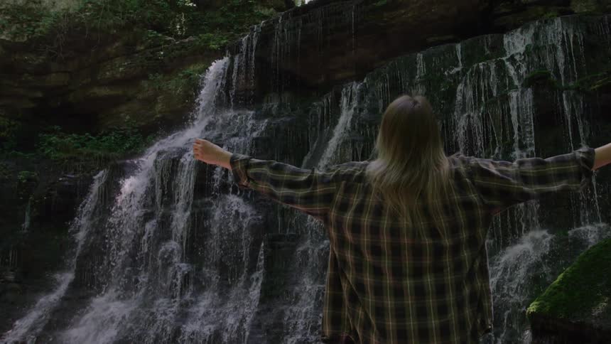 A woman raises her arms while standing in front of a waterfall