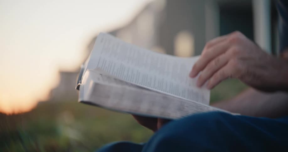 A persons hands slowly turn the pages of a book while sitting outside during a hazy sunset