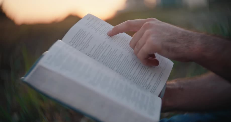 A mans hand points to a page in an open book while reading outdoors at sunset