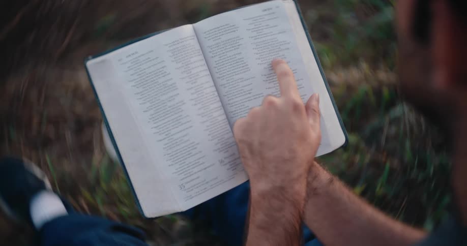 A high angle overhead view shows a man sitting in tall grass outdoors holding an open bible and using his finger to follow along while reading the text