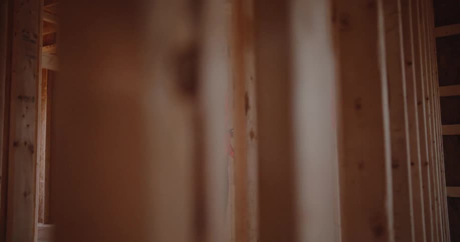 A construction worker in an orange safety vest and white hard hat holds a tablet and inspects areas inside a wood framed room