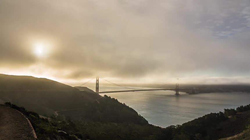 A time lapse view from a high vantage point shows the golden gate bridge shrouded in dense fog as the bright morning sun burns through the haze above the hills