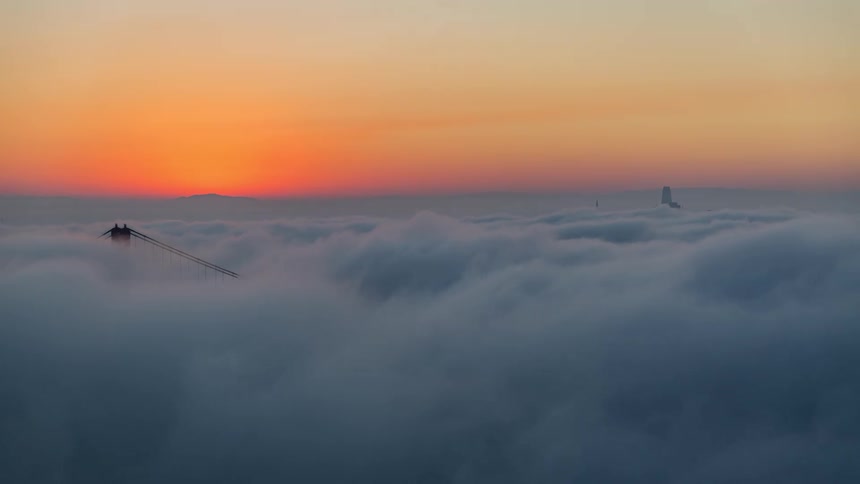 Bridge tower rises through dense clouds beneath an orange sunset