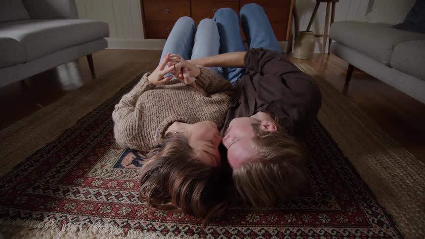 A medium view of a man and a woman in casual clothing stand on a wooden porch outside a house holding hands and smiling at each other in the morning light