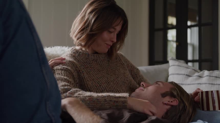 Man lying down on the lap of a woman sitting on a couch while she caresses him and both smile indoors during day time