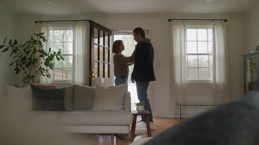 A man in a dark jacket and a woman in a sweater embrace and kiss affectionately while standing in the entryway of a brightly lit living room with a white sofa and large windows