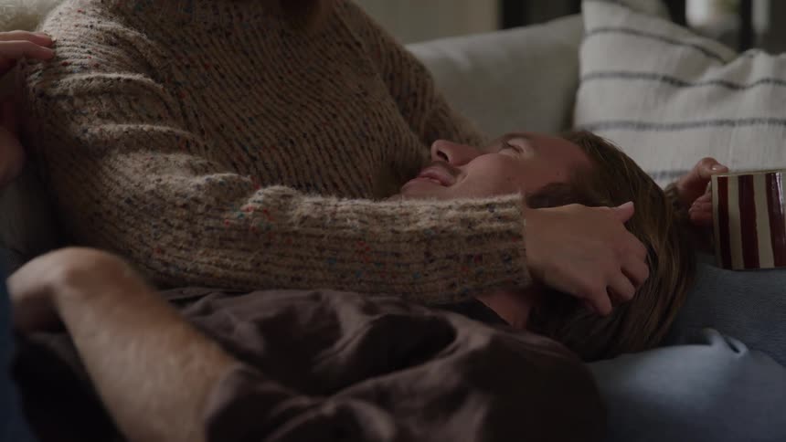 A man lies with his head in a womans lap on a sofa looking up at her face as the woman wearing a textured knit garment gently moves her hand over the mans hair