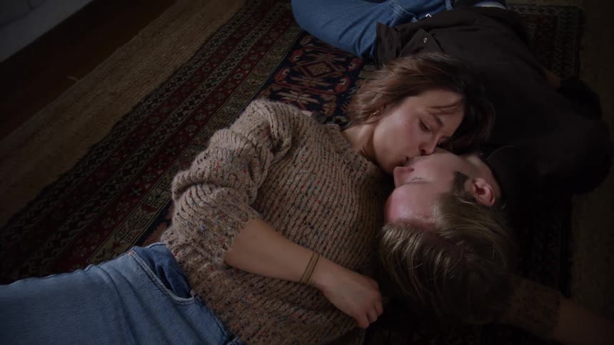 An overhead view of a man and a woman in sweaters and jeans lying on a colorful area rug embracing and kissing as they look sweetly at each other in a cozy intimate moment
