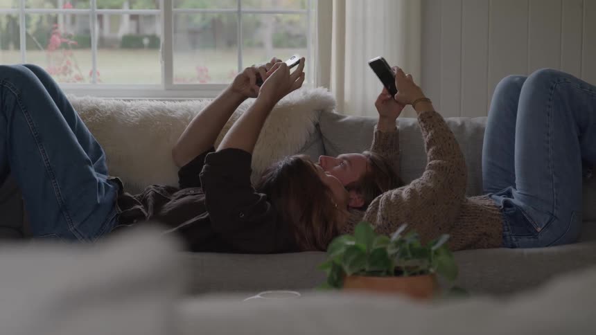 Two people lie on their backs on a couch facing opposite directions with their heads resting near the center holding mobile phones above their heads and actively using the devices while a window shows an outdoor view