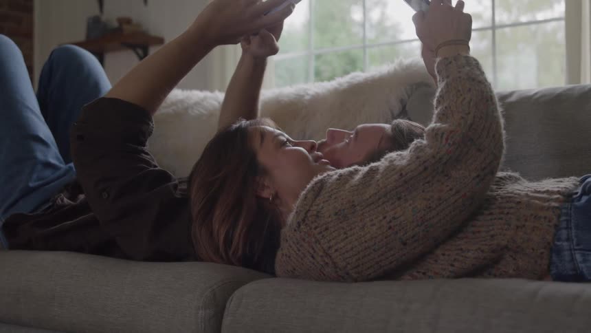 Close up of a young couple using their phones while lying together on a couch in a living room during daytime