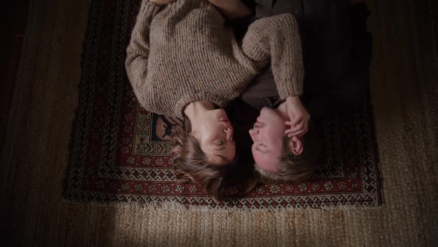 An overhead view shows a smiling man and woman lying on a colorful area rug facing each other with their heads touching as the woman gently strokes the mans head