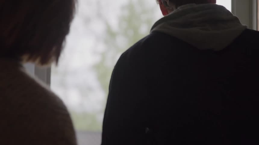 Man carrying a helmet in his hand kisses a woman in front of a door then walks outside the house into a lawn during day time