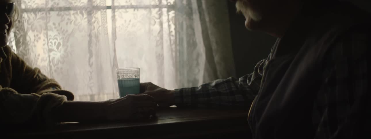 A man sits in a dark room handing a glass to someone