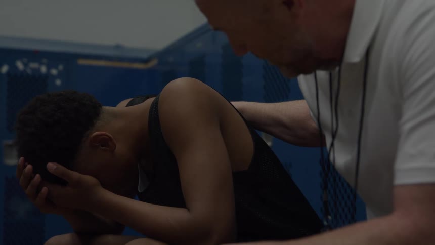 A coach in a white shirt sits beside a young man in a black tank top gently placing his hand on the players shoulder as the distressed player holds his head in his hands in a dimly lit blue locker room