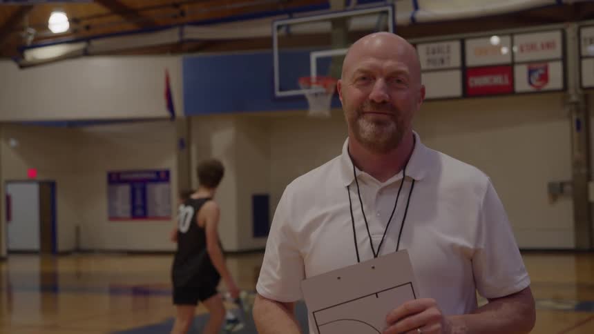 A bald man wearing a white collared shirt and a lanyard stands in the center foreground of a gymnasium looking directly at the camera with a smile and holding a clipboard that has a diagram on it while youth basketball players move and dribble in the back