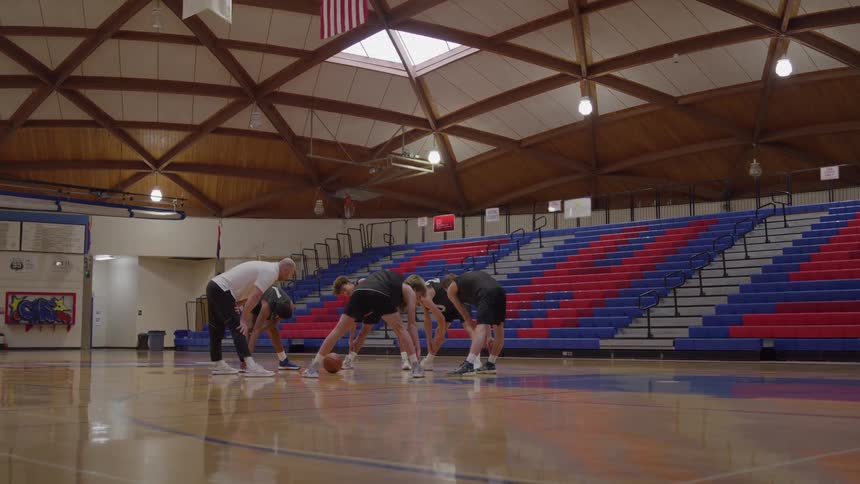 A group of four young men and one older man stand bent over at the waist with their hands near their feet on a glossy wood basketball court