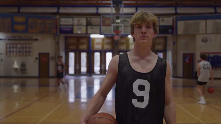 A confident young man with blonde hair and a black number nine basketball jersey holds a basketball and looks directly at the camera on a glossy gymnasium court with other players in the background