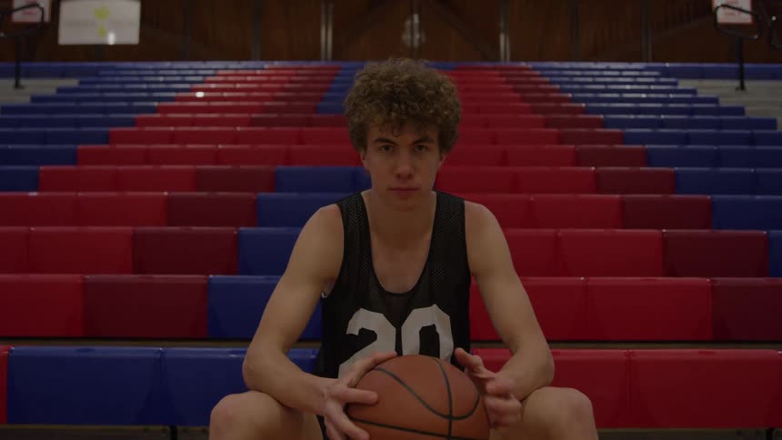 A young male basketball player wearing a dark numbered tank top sits facing the camera holding a brown basketball and in the center of a wide expanse of red and blue tiered gymnasium seating