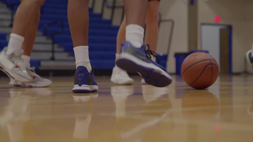 The lower legs and feet of multiple people wearing hightop athletic shoes and white socks shuffle and move on a reflective wood gymnasium floor with a basketball resting on the floor