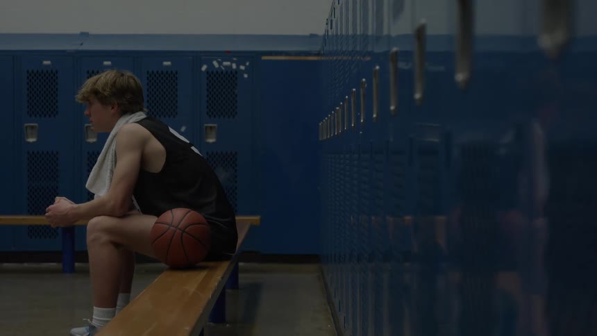 A side view of a young man with blonde hair wearing a black jersey and a white towel over his shoulder sits on a wooden bench with a basketball in a dimly lit blue locker room
