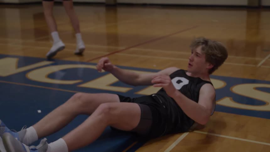 A young male basketball player wearing a dark jersey falls backward onto the wooden court floor then immediately extends his arm to grasp the hand of another player to receive assistance standing up before returning to the play area