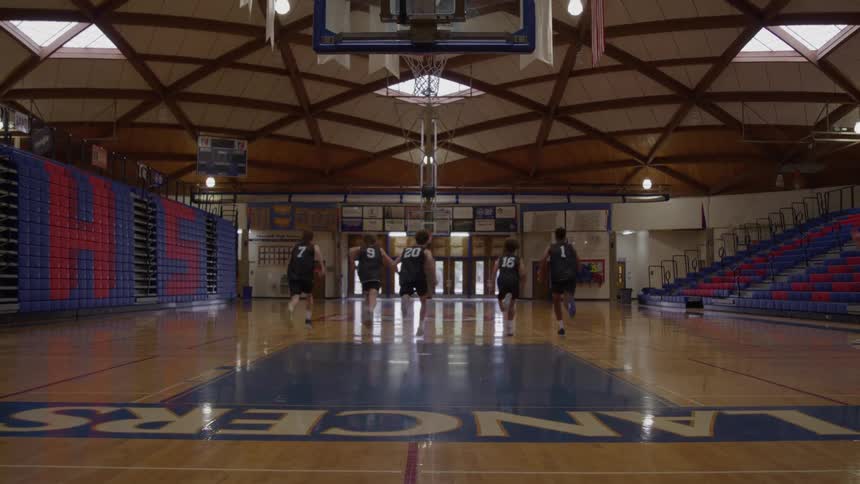 A group of five young male basketball players in black jerseys with numbers on their backs run suicides across a glossy wooden gymnasium court toward the camera which is positioned low near the basketball net