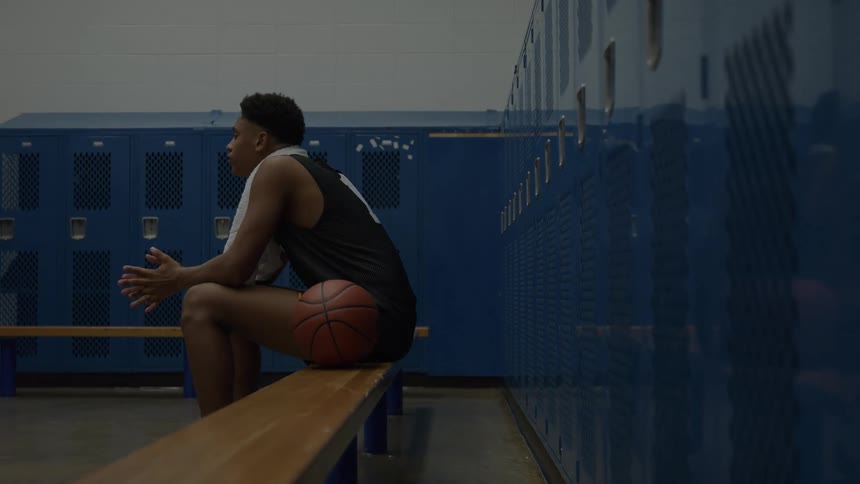 A basketball player with a towel around his neck sits on a locker room bench with the ball resting against his leg looking thoughtful and tense