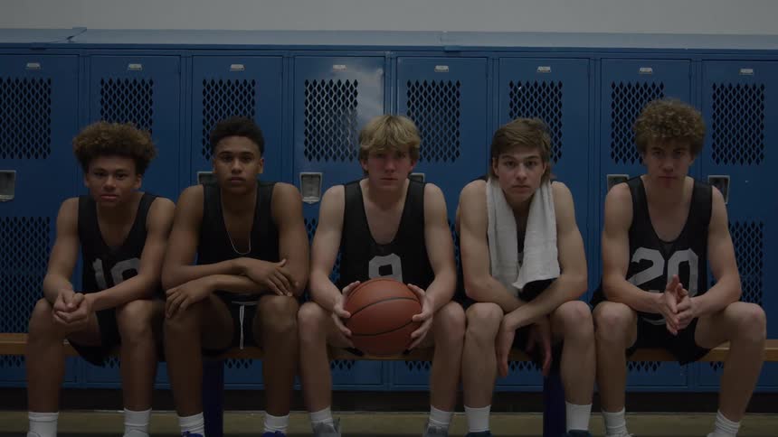 Five young male basketball players in black jerseys sit quietly side by side on a wooden bench against a wall of blue lockers with one holding a basketball and three looking up with serious expressions