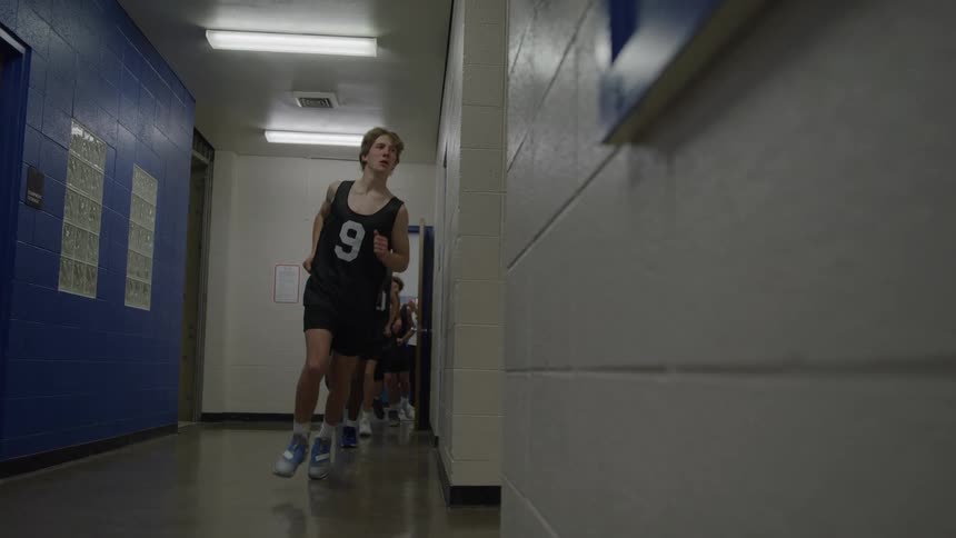 A group of people wearing dark numbered sports jerseys and shorts run toward the camera down a brightly lit indoor hallway with white and blue walls emerging from an open doorway at the end of the hall