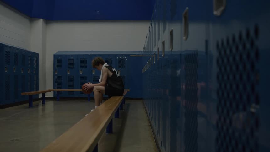 A young male athlete sits alone on a locker room bench holding a basketball and staring down in quiet contemplation