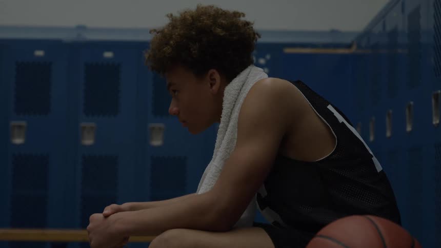 A basketball player with a towel around his neck sits on a locker room bench with head bowed beside a ball looking dejected